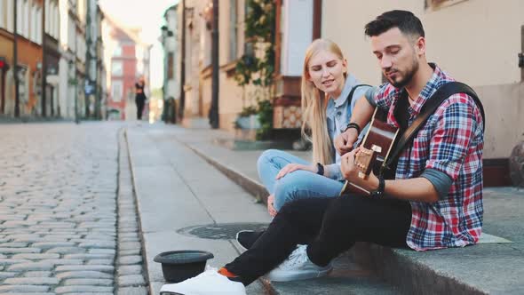 Young Couple of Street Singers Sitting on Sidewalk Playing Guitar and Singing alt