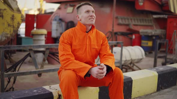 Young Man in Orange Uniform Sitting During His Break By the Sea in the Harbor alt