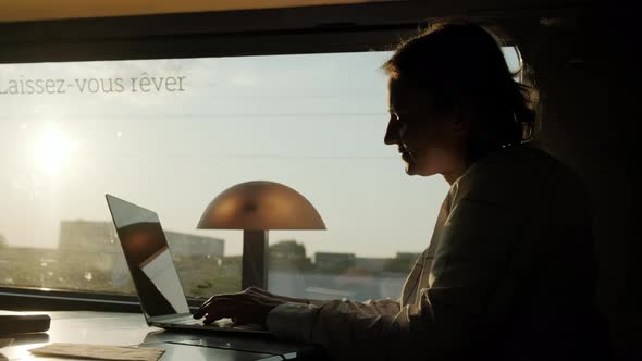 Silhouette of Woman Works on Laptop While Riding Train Sitting Near Window on Sunny Day alt