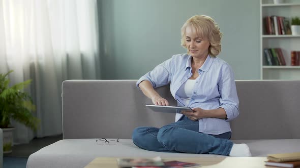 Good-Looking Blonde Senior Woman Sitting on Sofa and Scrolling on Tablet, Apps alt
