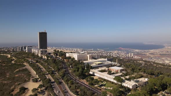 Flight to Haifa city around a tall building on a hill surrounded by trees and roads, with the harbor alt
