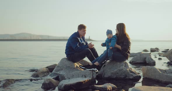 Husband and Wife are Sitting on the Rocks Near the Water and Playing with Their Young Son alt