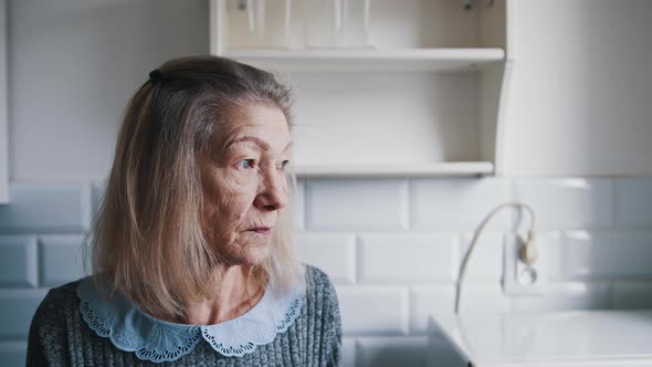 Portrait of Elderly Gray Haired Lady Looking Through the Kitchen Window and Thinking alt