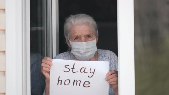 An Old Gray-haired Woman in a Medical Mask Stands at Home Near the Window and Holds a Poster with