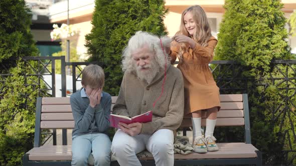 Pretty Caucasian Girl Making Ponytails on Long Grey Hair of Grandfather While Senior Man Reading alt