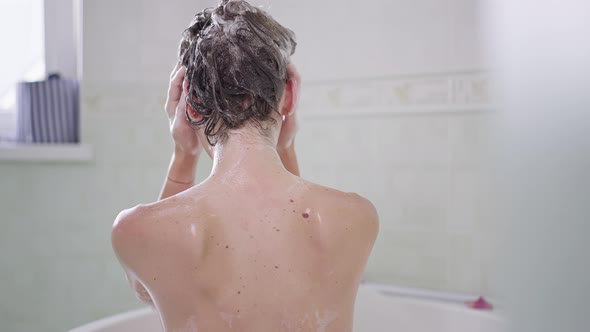 Back View of Young Slim Woman Shampooing Hair Sitting in Bath at Home alt