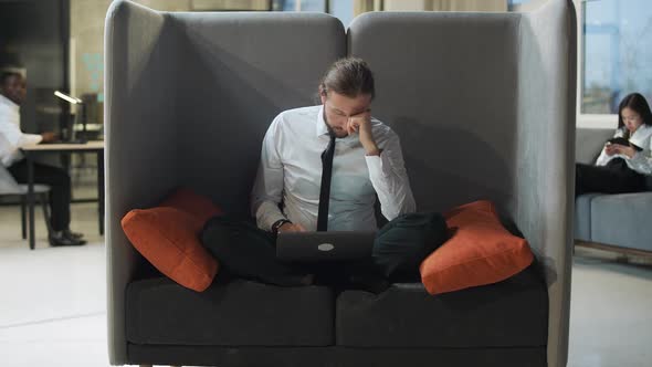 Office Worker Sits on a Large Chair and Works at a Laptop Office Work in an Open Space a Tired Man alt