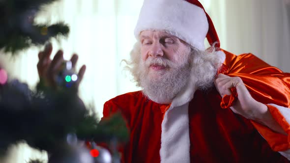 Bearded Senior Caucasian Greyhaired Man with Blue Eyes in Santa Clause Costume Standing at Decorated alt