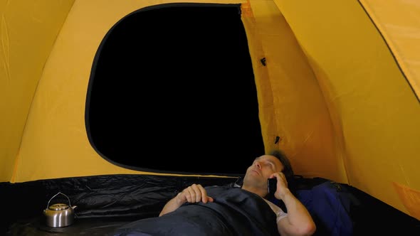 Tourist Resting in Sleeping Bag inside the Tent and Talking by Smartphone, Alpha in