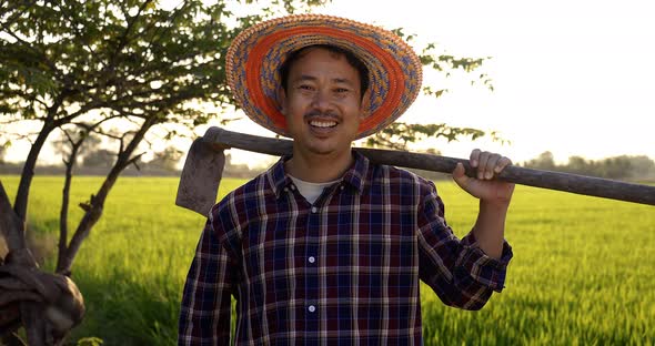 Slow motion Happy Asian Farmer holding a hoe and looking at camera green paddy field on background. alt