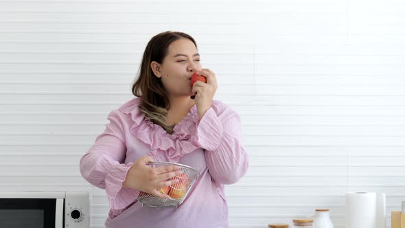 Happy fat woman standing and eating apple deliciously in kitchen alt