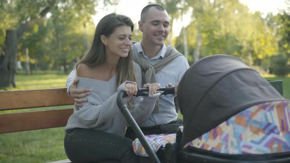 Happy Couple of Young Caucasian Parents Rocking Baby Carriage, Hugging and Talking. Portrait of alt