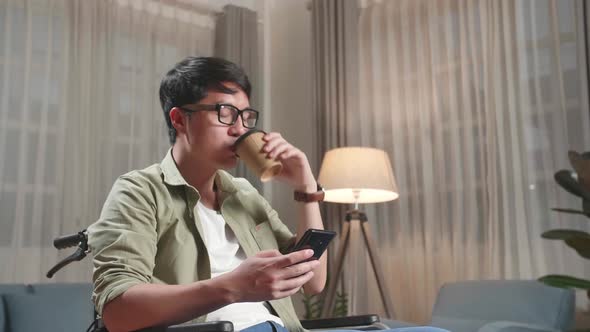 Asian Man Sitting In A Wheelchair While Use Mobile Phone And Drinking Coffee At Home alt