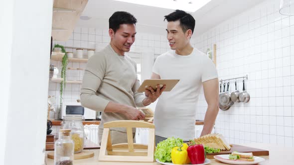 Asia gay couple using tablet and preparing the breakfast, sandwich vegetable on table.
