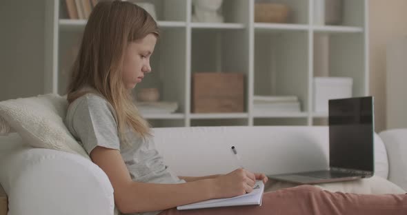 Teen Girl Is Doing Homework in Room of Flat After School, Writing in Exercise Book, Sitting on Couch