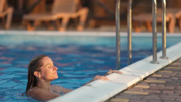 Woman Swims in a Pool with Clear Water on the Background of a Summer Sunset on Vacation alt