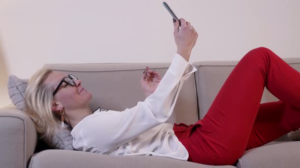 A Cheerful Blonde Woman with Glasses is in the Room Lying on the Sofa and Looking at the Phone alt