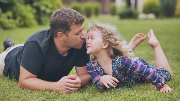 A Father Kisses His Young Daughter in the Garden While Lying on the Grass alt