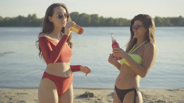 Side View of Two Young Caucasian Women Clinking Bottles with Alcohol on Summer Beach and Drinking alt