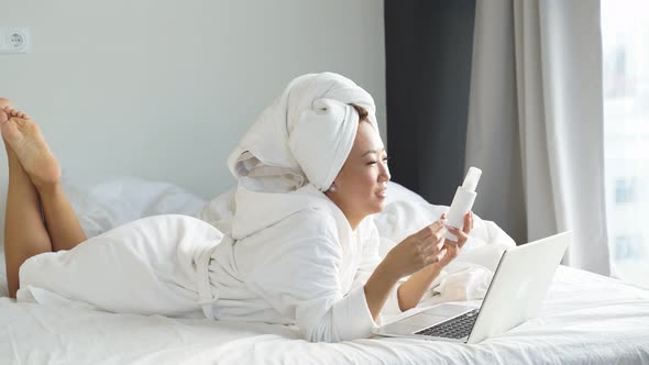 Attractive Girl Lying on the Bed Talking on a Video Link on a Laptop and Showing Body Cream Face alt