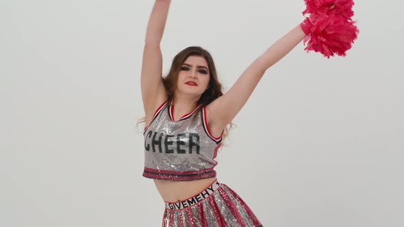 Young Cheerleader with Red Pompoms in Uniform is Dancing on White Background in Studio alt