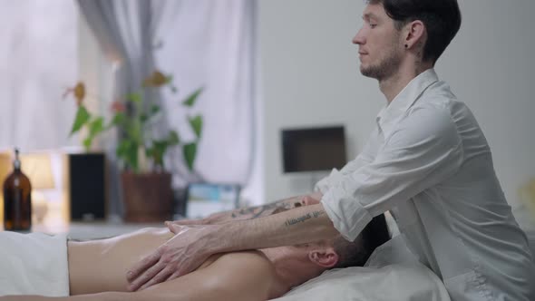 Side View of Concentrated Male Masseur Massaging Chest of Bearded Relaxed Man in Spa Salon alt