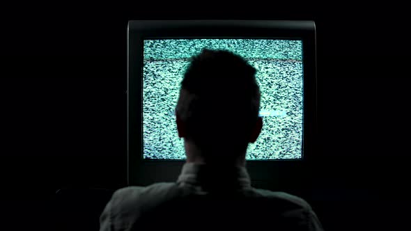 Rear View of a Silhouette of a Man Sitting in Front of a Flickering TV in a Dark Studio on a Black alt