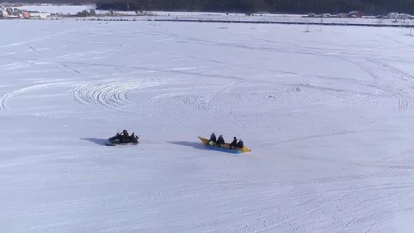 Aerial View Happy people enjoying banana boat ride tied to a snowmobile alt