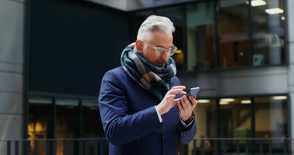 A Grayhaired Man Uses a Mobile Phone Standing in Business Center of the City alt