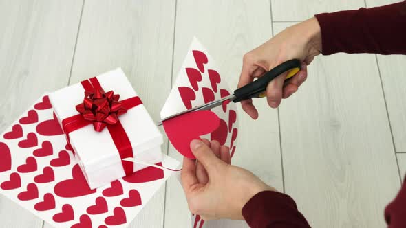 Woman hands cutting out red heart shape from paper cardboard using scissors.