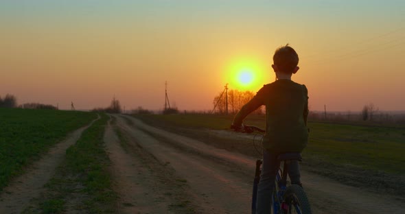 A Boy Is Riding a Bicycle on a Pathway at Sunset. Filming From the Back alt