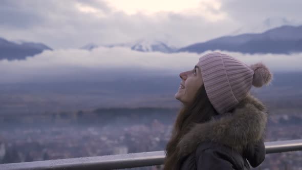 Smiling Woman Enjoying the View of City Under Misty Snowy Mountains alt