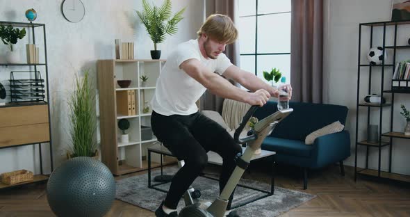 Guy in Sportswear Hard Cycling on Stationary Bike During Morning Home Workout alt