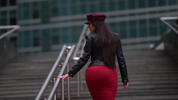 a Brunette with Long Hair in a Black Leather Jacket a Red Skirt and a Burgundy Cap Climbs the Stairs alt