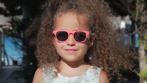 Close-up Portrait of a Beautiful Little Girl in Pink Glasses, Cute Smiling, Looking at the Camera alt