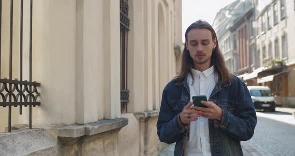 Young Long Haired Guy Using His Smartphone and Looking Forward at Old City Street, Bearded Hipster alt