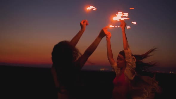 Young Women Having Fun Dancing with Arms Outstretched and Celebrating with Sparklers at Beach Party
