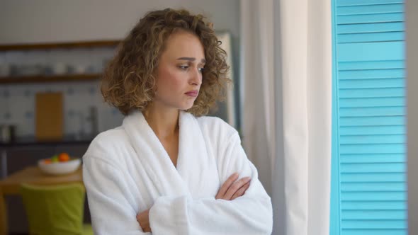 Sad Young Woman in Bathrobe Standing at Window and Looking Into Distance alt