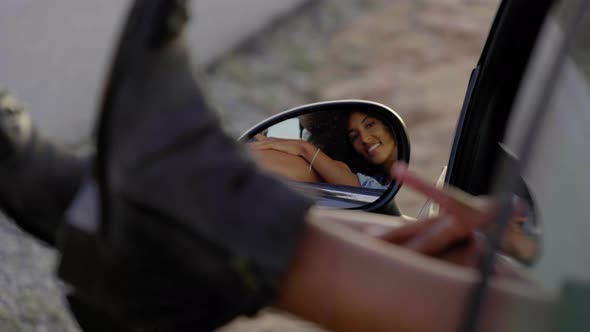Mirroring of young woman during road trip, leaning feet out of window alt