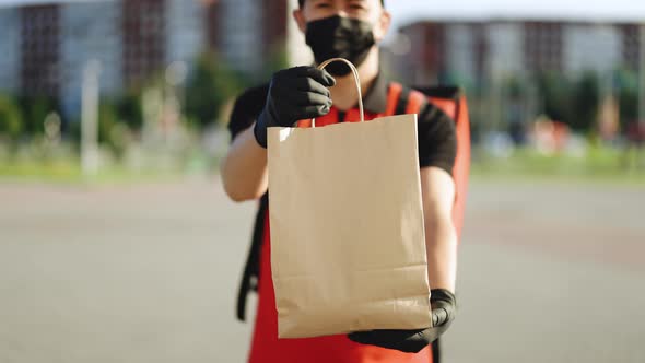 Deliveryman in Protective Medical Face Mask Gloves Holding Paper Bag With Food at Street Outdoors