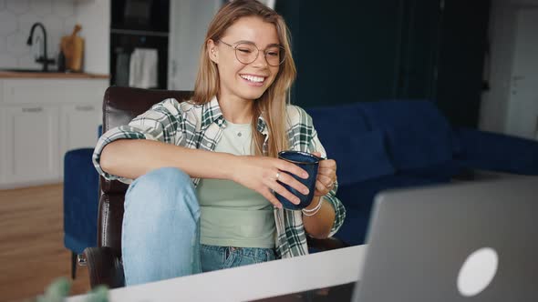 Young Cheerful Woman Sitting at Home with Cup of Tea and Watching Movie Online on Laptop alt
