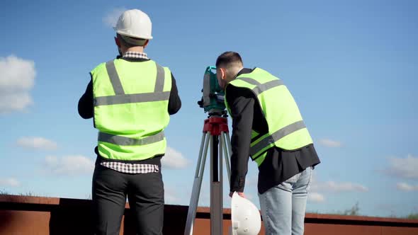 Back View Two Male Caucasian Architects with Theodolite and Binoculars Looking Away Talking Standing alt