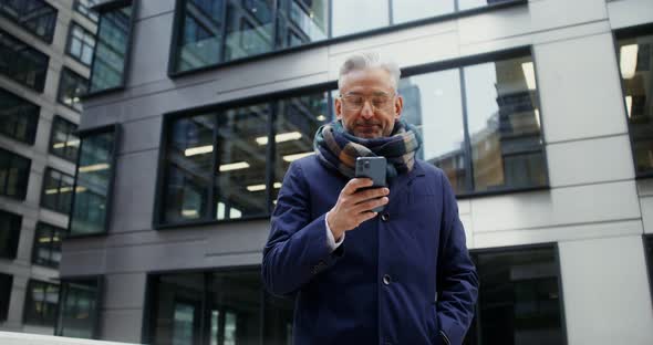 A Grayhaired Man Uses a Mobile Phone Standing in Business Center of the City alt