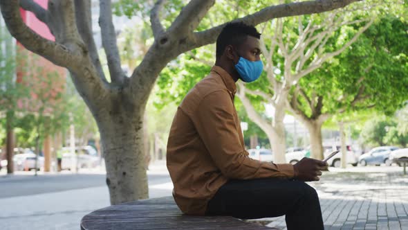African american businessman wearing face mask using tablet sitting on bench alt