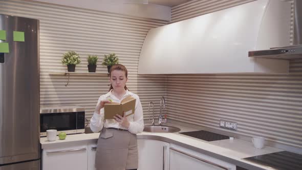 Hipster Young Woman Search a Recipe in Her Cookbook While Standing in the Kitchen