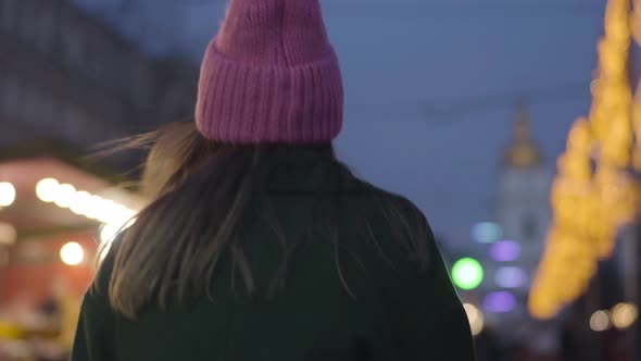 Camera Following Young Caucasian Woman in Pink Hat and Green Coat Walking To Children's Carousels on alt