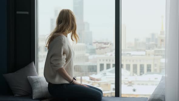 a woman sits on the windowsill and looks out the window at the city