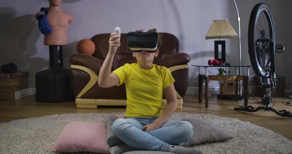 Young Caucasian Teen with Curly Hair in VR Headset Holding Remote Control and Looking Around. Child alt