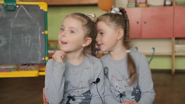 Twin Girls Sitting on a Chair Answers Questions. Girls Give Interviews in a Classroom Against the