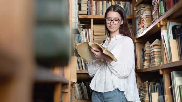Young Attractive Student in Glasses Turns Pages in the Book Which She Took From Books on Shelves in alt
